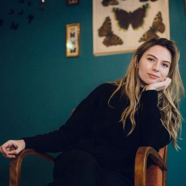 Woman with long curly blonde hair wearing black jumper sits in armchair and rests her face in her hand